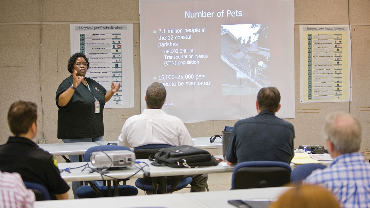 Woman speaker standing next to a large screen presenting to a group of people